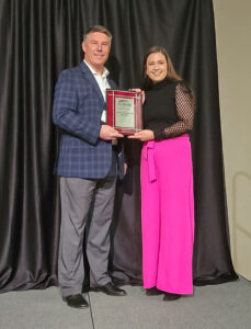 Sarah Crawford and Ted Rogalski stand on stage holding a plaque