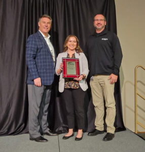 Tim Heitzig, Amy Knox and Ted Rogalski stand on a stage holding a plaque
