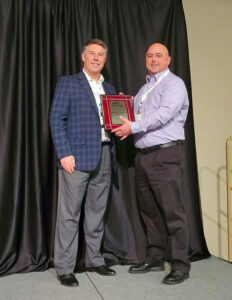 Patrick Small stands on a stage with Ted Rogalski holding a plaque